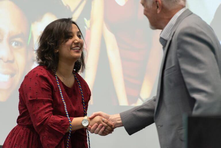 Smiling Latina student in red dress shakes hand over older man in suit jacket