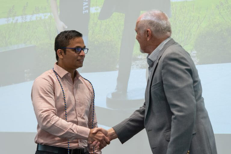 male student with dark hair and glasses in colored shirt shaking hands with older man in suit in front of screen projecting UIS campus