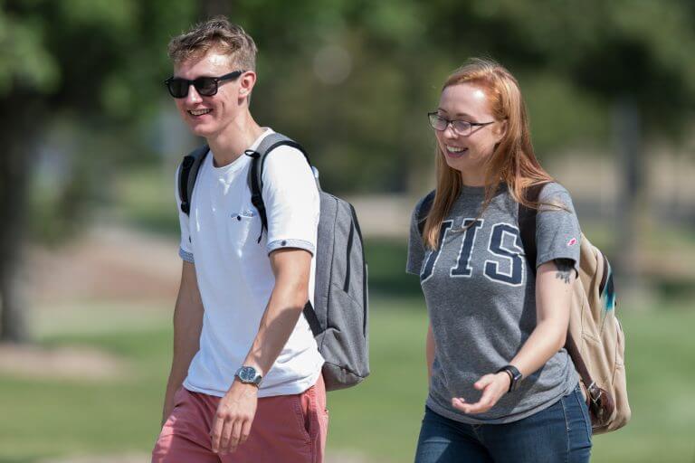 Male student in sunglasses wearing backpack and female student in glasses and UIS t-shirt with backpack walking on campus