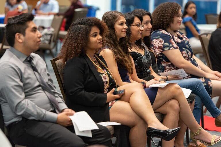 47754703452_bd23c4e9a7_o-768x513.jpg Diverse students of color in formal wear sit in a row listening to speaker